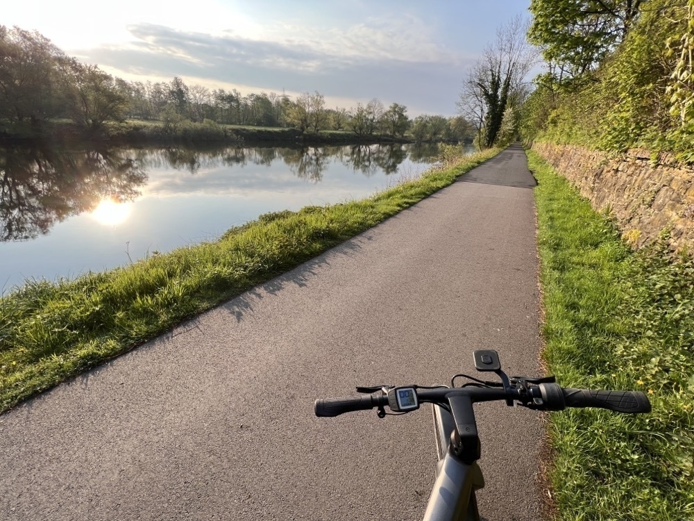 Front part of an e-bike in the foreground with a river in the background. The sun is reflected in the water. 